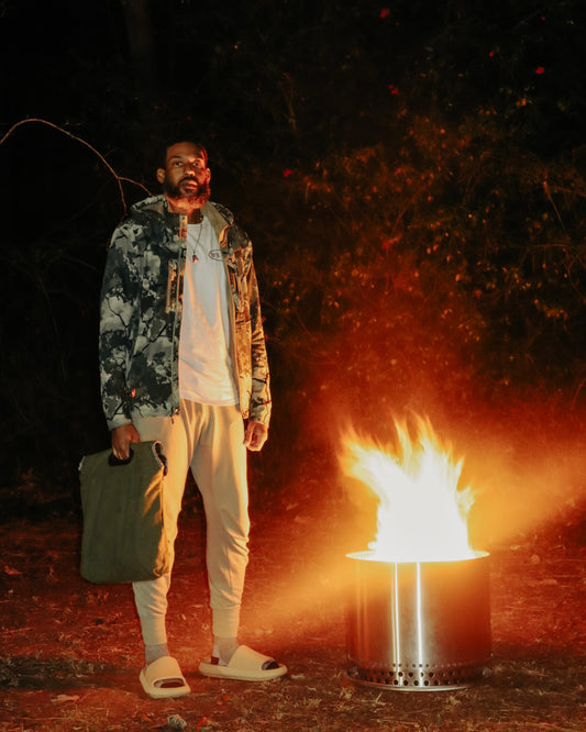 Man standing by a campfire at night, wearing a camouflage jacket and carrying a tote bag, with a serene expression on his face. The fire casts a warm glow on the surrounding foliage, enhancing the outdoor nocturnal atmosphere.
